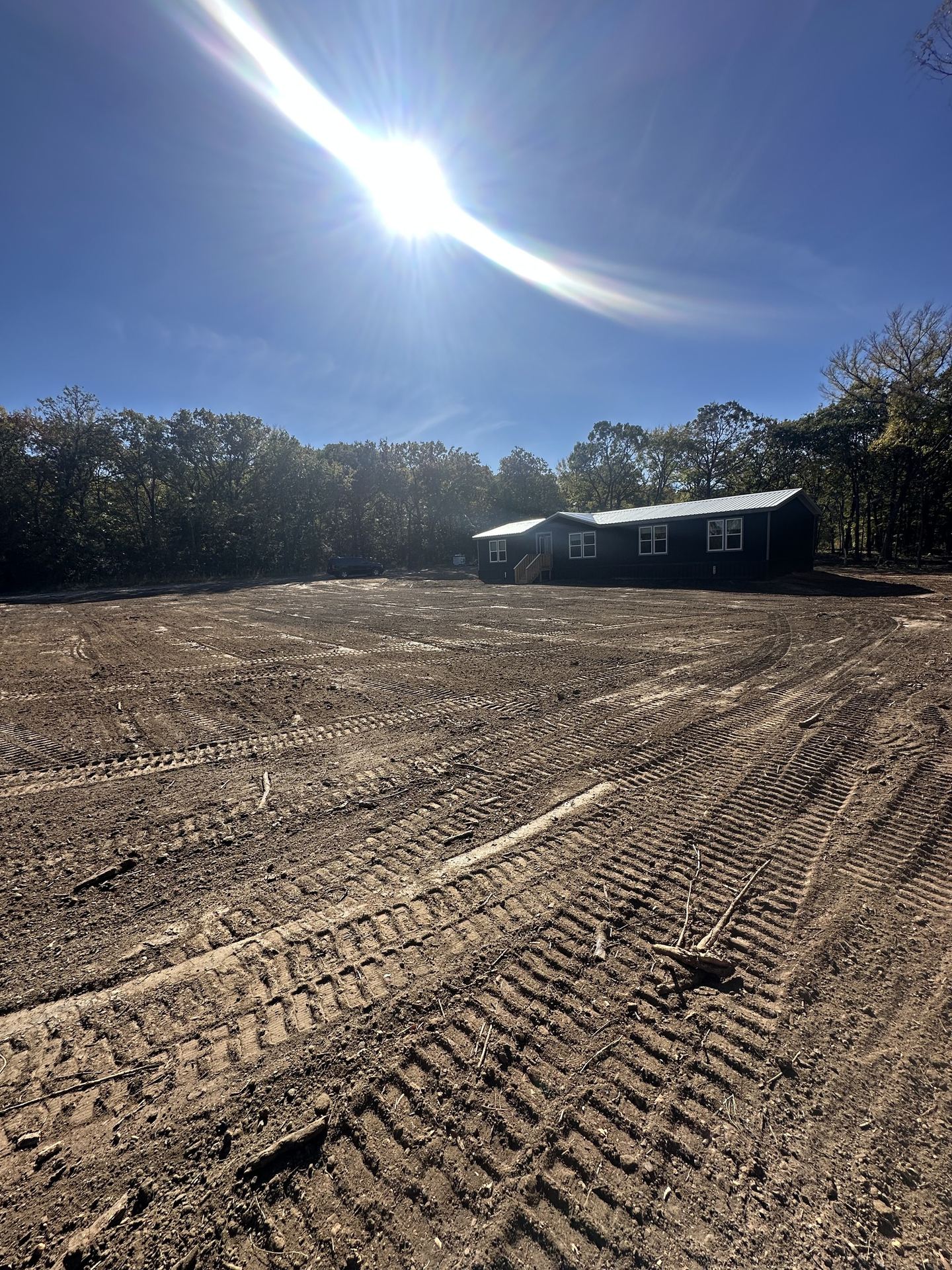 Cleared and graded lot with dozer tracks, a new house at the tree line and the sun overhead