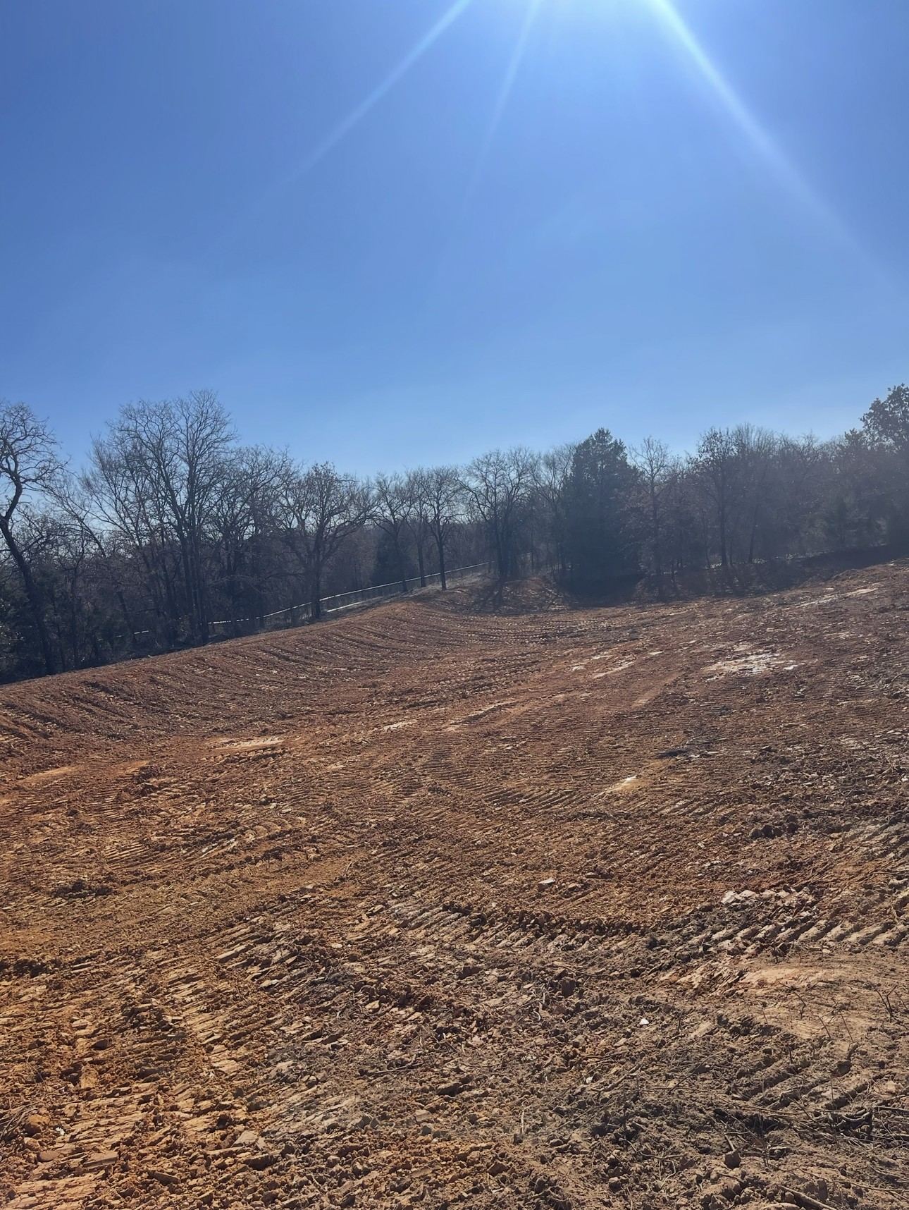Hillside cleared down to red Oklahoma dirt with dozer tracks and a tree line beyond