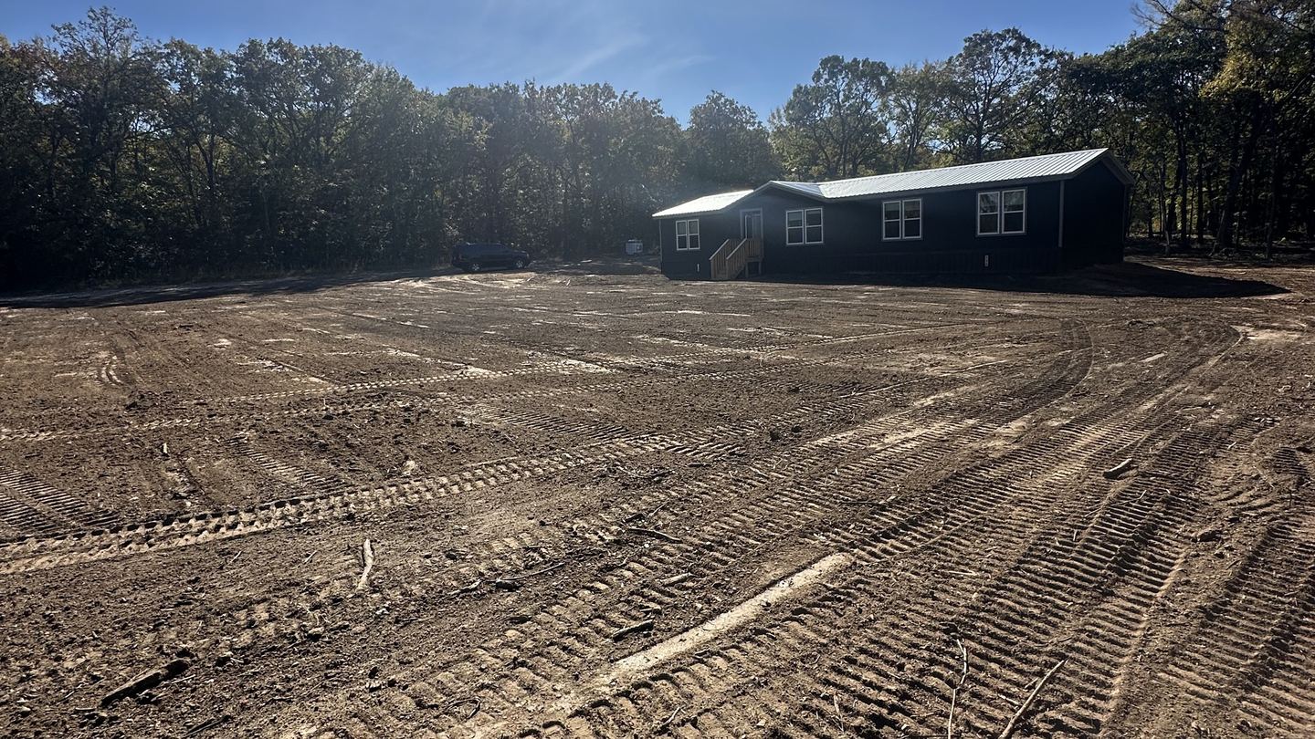 Freshly cleared and graded lot with dozer tracks across the dirt, a tree line and a new house behind